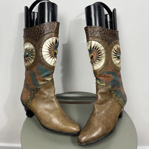May include: A pair of brown leather boots with decorative detailing. The boots feature a mix of textures, including patterned leather and embellishments. Each boot has a heel and a decorative circular design with a gemstone in the center.