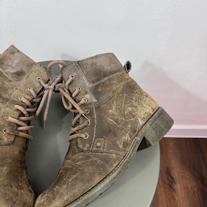 K&ouml;nnte beinhalten: Ein Paar kn&ouml;chelhohe Schn&uuml;rstiefel aus abgenutztem braunem Leder mit abgerundeter Zehenpartie, niedrigem Blockabsatz und passenden braunen Schn&uuml;rsenkeln, die durch messingfarbene Metall&ouml;sen gef&uuml;hrt werden. Die Stiefel haben eine texturierte Optik.