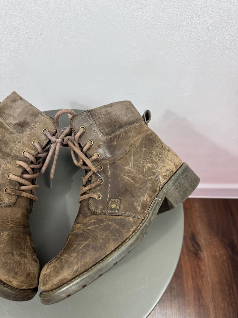 K&ouml;nnte beinhalten: Ein Paar kn&ouml;chelhohe Schn&uuml;rstiefel aus abgenutztem braunem Leder mit abgerundeter Zehenpartie, niedrigem Blockabsatz und passenden braunen Schn&uuml;rsenkeln, die durch messingfarbene Metall&ouml;sen gef&uuml;hrt werden. Die Stiefel haben eine texturierte Optik.