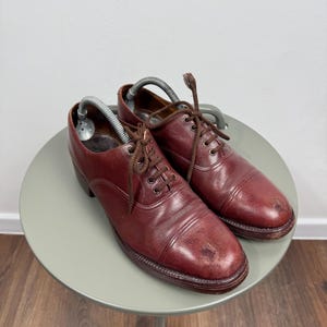 May include: A pair of burgundy leather dress shoes with brown laces. The shoes are on a small, round, light green table. Shoe trees are inserted in the shoes. The shoes show signs of wear.