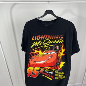Peut inclure: T-shirt noir avec un motif de Flash McQueen du film Cars. Le design comprend le texte "Lightning McQueen", le num&eacute;ro "95" et les phrases "Rule the Track", "The Champ", "The Myth" et "The Legend".