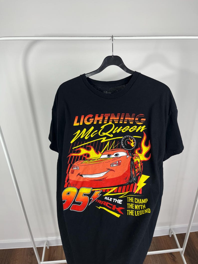 Peut inclure: T-shirt noir avec un motif de Flash McQueen du film Cars. Le design comprend le texte "Lightning McQueen", le num&eacute;ro "95" et les phrases "Rule the Track", "The Champ", "The Myth" et "The Legend".