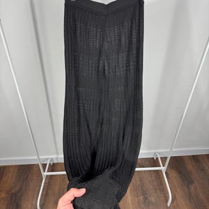May include: Black, ribbed knit pants hanging on a clothing rack. The pants have a wide leg and a textured appearance. The fabric appears to be lightweight and suitable for warmer weather. The pants are displayed against a neutral background.