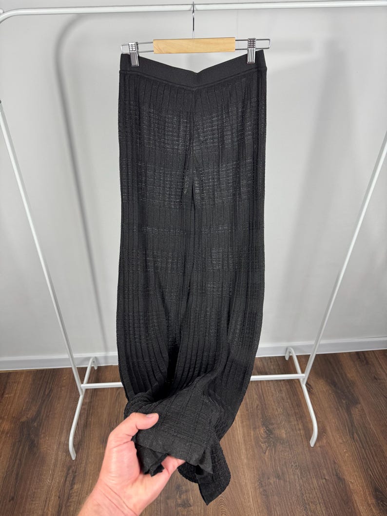 May include: Black, ribbed knit pants hanging on a clothing rack. The pants have a wide leg and a textured appearance. The fabric appears to be lightweight and suitable for warmer weather. The pants are displayed against a neutral background.