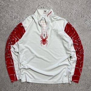 May include: White athletic top with red spiderweb pattern on the sleeves. A red spider graphic is on the chest. The top has a mock neck with a zip and the brand name "Spyder" in red.