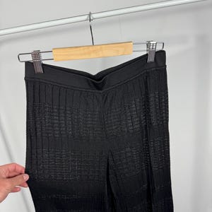 May include: Black textured knit pants hanging on a wooden hanger. The pants have a high waist and a vertical ribbed pattern. The fabric appears to have a subtle sheen. The pants are displayed against a plain white background.