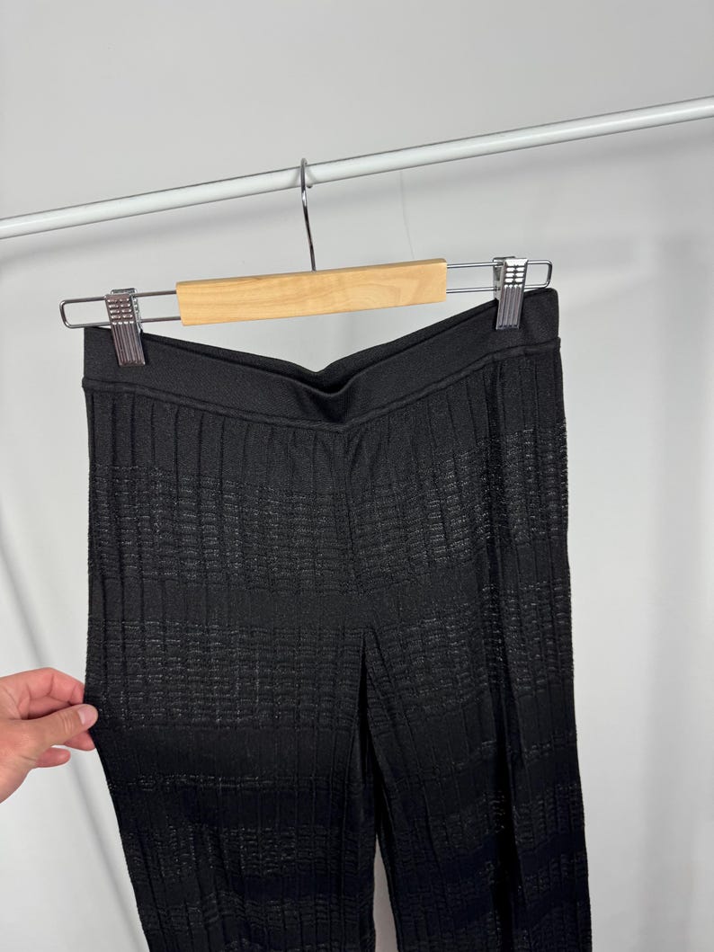 May include: Black textured knit pants hanging on a wooden hanger. The pants have a high waist and a vertical ribbed pattern. The fabric appears to have a subtle sheen. The pants are displayed against a plain white background.
