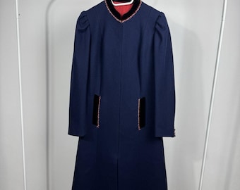 Puede incluir: Un abrigo largo de color azul marino con cuello alto, ribete de terciopelo negro y costuras decorativas rojas. El abrigo incluye dos bolsillos verticales con detalles de terciopelo negro y costuras rojas, mangas largas y un forro interior rojo.