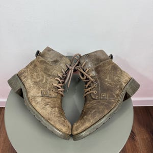 K&ouml;nnte beinhalten: Ein Paar kn&ouml;chelhohe Stiefel aus abgenutztem braunem Leder mit Schn&uuml;rung, passenden braunen Schn&uuml;rsenkeln, abgerundeter Zehenpartie, niedrigem Blockabsatz und einer Zugschlaufe am Schaft. Die Stiefel weisen sichtbare Kratzer und eine Textur auf.