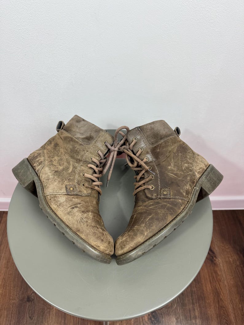 K&ouml;nnte beinhalten: Ein Paar kn&ouml;chelhohe Stiefel aus abgenutztem braunem Leder mit Schn&uuml;rung, passenden braunen Schn&uuml;rsenkeln, abgerundeter Zehenpartie, niedrigem Blockabsatz und einer Zugschlaufe am Schaft. Die Stiefel weisen sichtbare Kratzer und eine Textur auf.