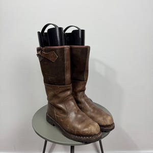 May include: A pair of mid-calf boots featuring a combination of distressed brown leather on the lower section and dark brown suede on the upper shaft. Each boot includes a side strap with a buckle and black plastic boot trees inserted inside.