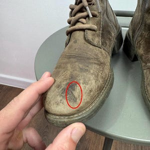 K&ouml;nnte beinhalten: Ein Paar geschn&uuml;rte Stiefel aus abgenutztem olivbraunem Leder mit abgerundeter Zehenpartie, passenden braunen Schn&uuml;rsenkeln und einem sichtbaren Kratzer auf der Zehenkappe des linken Stiefels, der durch einen d&uuml;nnen roten Ovalrahmen markiert ist.