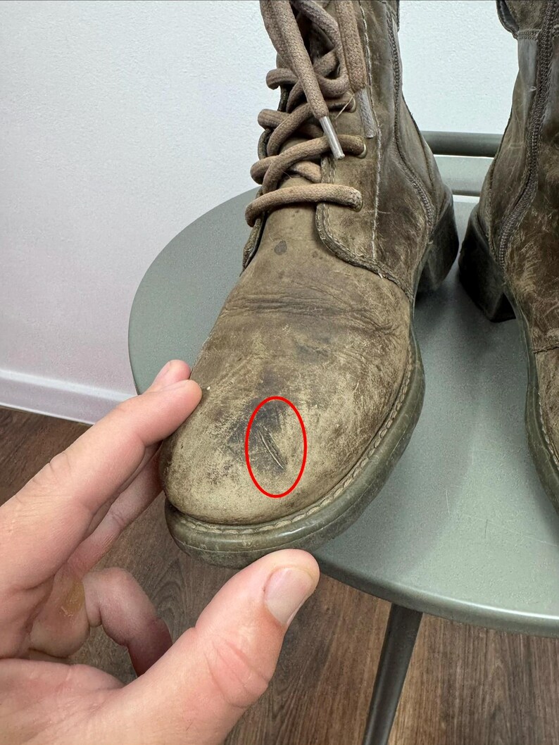 K&ouml;nnte beinhalten: Ein Paar geschn&uuml;rte Stiefel aus abgenutztem olivbraunem Leder mit abgerundeter Zehenpartie, passenden braunen Schn&uuml;rsenkeln und einem sichtbaren Kratzer auf der Zehenkappe des linken Stiefels, der durch einen d&uuml;nnen roten Ovalrahmen markiert ist.