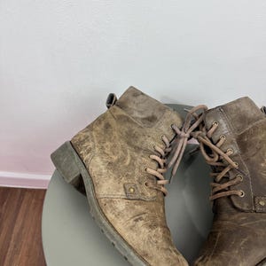 K&ouml;nnte beinhalten: Ein Paar kn&ouml;chelhohe Stiefel aus abgenutztem beigefarbenem Leder mit Schn&uuml;rung, passenden Schn&uuml;rsenkeln, Metall&ouml;sen, einem seitlichen Nietendetail, einer Zugschlaufe an der Ferse und einem niedrigen Blockabsatz mit profilierter Gummisohle.