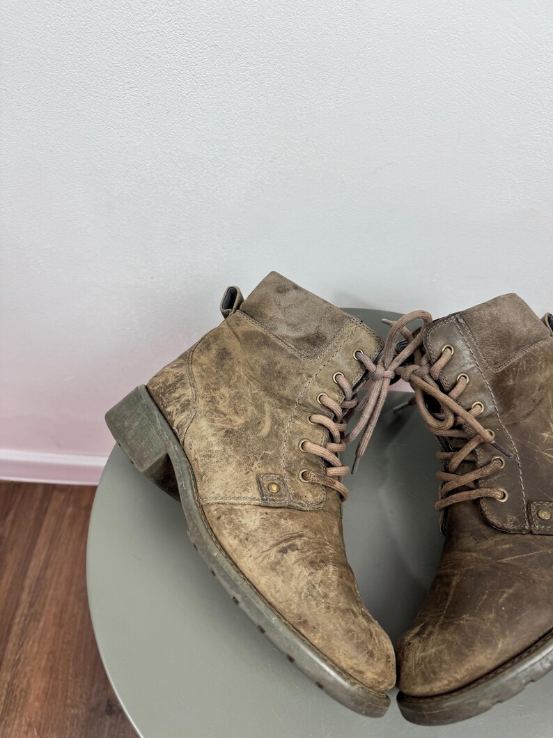 K&ouml;nnte beinhalten: Ein Paar kn&ouml;chelhohe Stiefel aus abgenutztem beigefarbenem Leder mit Schn&uuml;rung, passenden Schn&uuml;rsenkeln, Metall&ouml;sen, einem seitlichen Nietendetail, einer Zugschlaufe an der Ferse und einem niedrigen Blockabsatz mit profilierter Gummisohle.