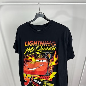 Peut inclure: T-shirt noir avec un motif de Flash McQueen du film Disney Cars. Le graphisme comprend McQueen, le num&eacute;ro 95, des flammes et le texte "Rule the Track" et "The Champ, The Myth, The Legend."