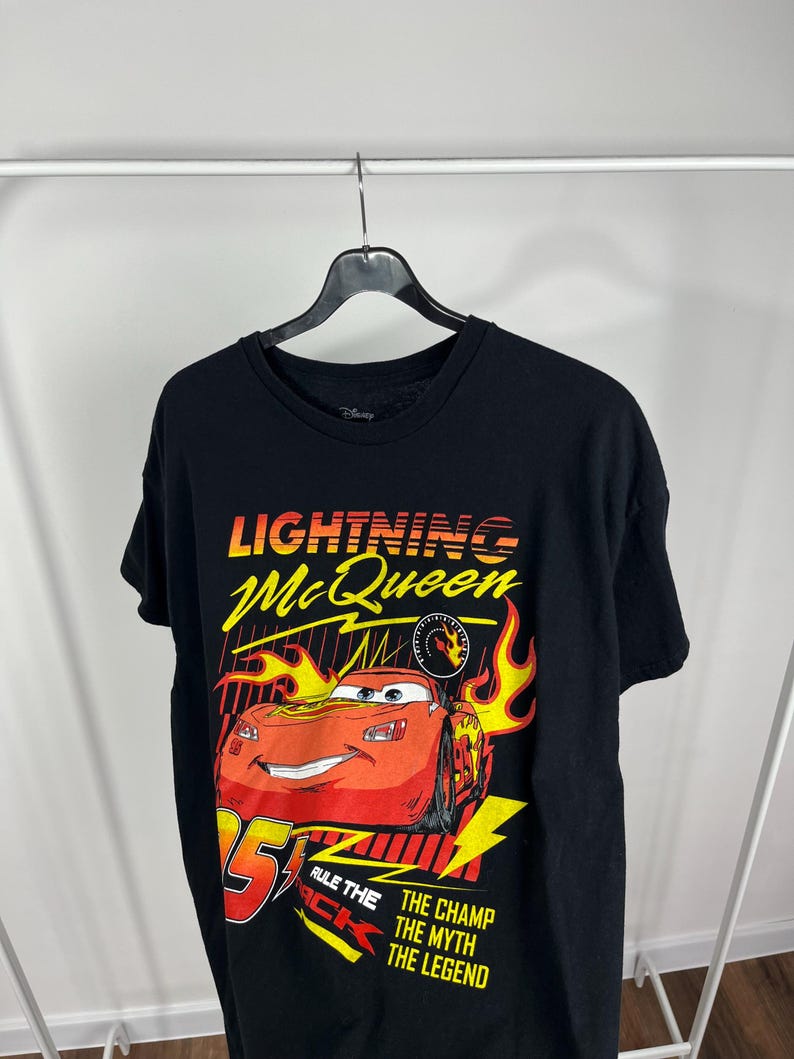 Peut inclure: T-shirt noir avec un motif de Flash McQueen du film Disney Cars. Le graphisme comprend McQueen, le num&eacute;ro 95, des flammes et le texte "Rule the Track" et "The Champ, The Myth, The Legend."