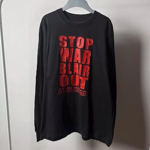 Puede incluir: Camiseta negra de manga larga con el texto rojo "STOP WAR BLAIR OUT" impreso en la parte delantera. La camiseta está colgada en una percha metálica. El texto incluye una dirección web en la parte inferior.