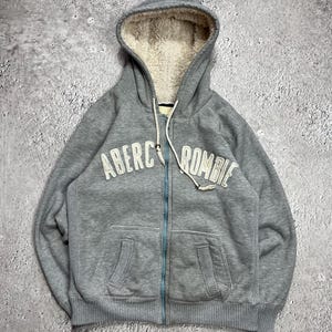 May include: A grey zip-up hoodie with a white sherpa-lined hood. The hoodie features the text "Abercrombie" in white block letters across the chest. It has a light blue zip and two front pockets. The fabric appears to be a soft, comfortable material.