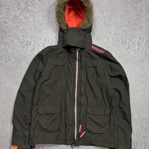 May include: A dark olive green hooded jacket featuring a bright orange interior lining and a faux fur trim on the hood. The jacket includes a front zipper closure, four flap pockets, and the text Superdry printed in red on the left shoulder.