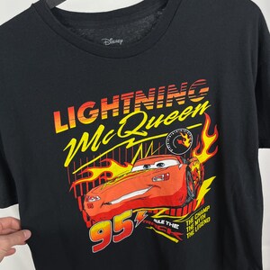Peut inclure: T-shirt noir avec un motif de Flash McQueen du film Cars de Disney. Le graphisme comprend le texte "Lightning McQueen" en jaune et orange, une voiture de course rouge et le num&eacute;ro "95".