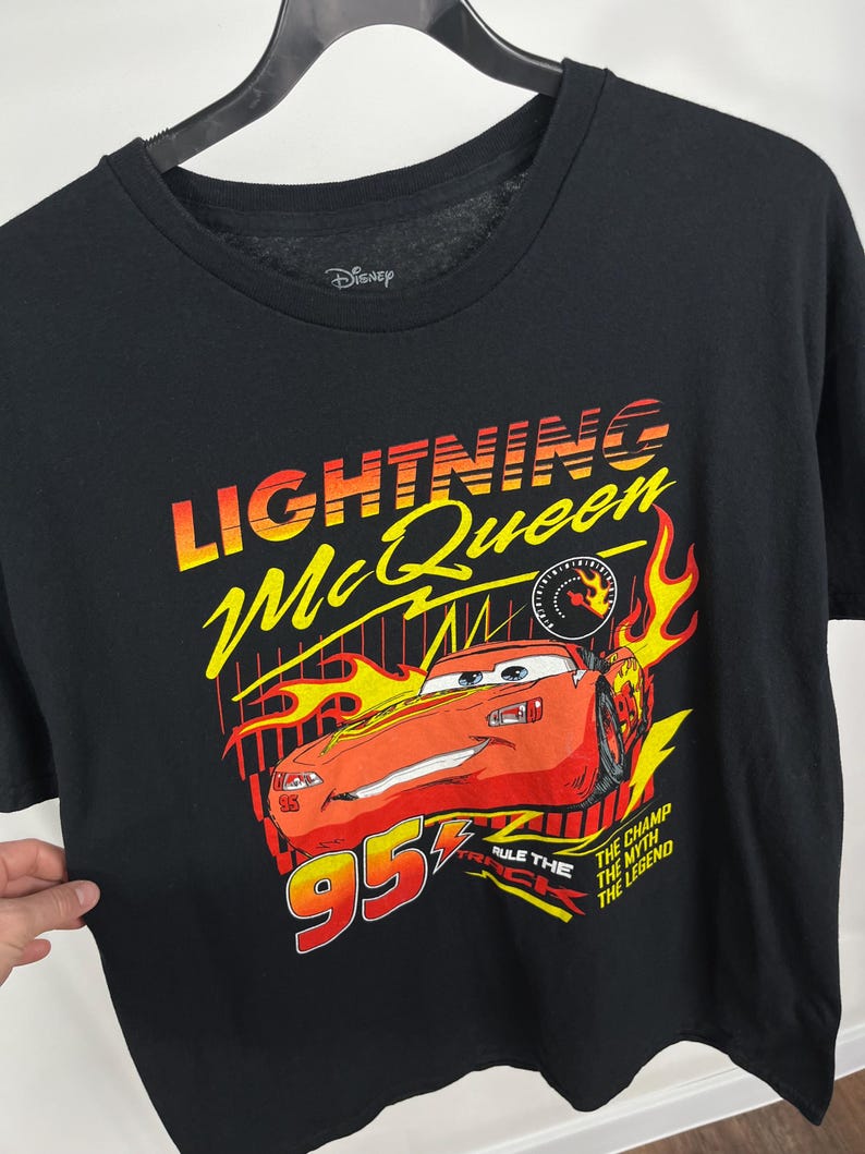 Peut inclure: T-shirt noir avec un motif de Flash McQueen du film Cars de Disney. Le graphisme comprend le texte "Lightning McQueen" en jaune et orange, une voiture de course rouge et le num&eacute;ro "95".