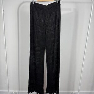 May include: Black, wide-leg pants with a textured, ribbed design hanging on a white clothing rack. The pants have a high waist and a slight flare at the hem. The fabric appears to be lightweight and semi-sheer.