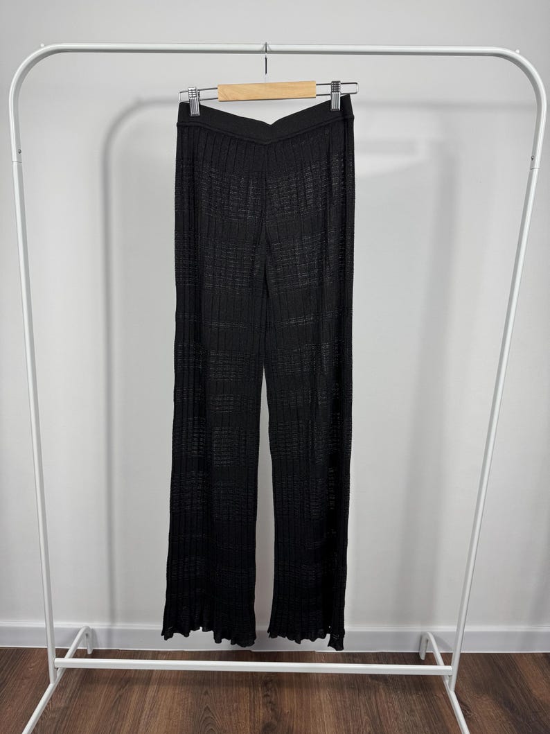 May include: Black, wide-leg pants with a textured, ribbed design hanging on a white clothing rack. The pants have a high waist and a slight flare at the hem. The fabric appears to be lightweight and semi-sheer.