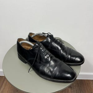 May include: A pair of black leather dress shoes with a classic cap-toe design. The shoes are laced up and sit on a small, round, grey table. The shoes are in good condition, with some visible wear.