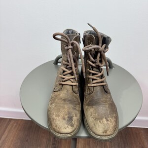 K&ouml;nnte beinhalten: Ein Paar abgenutzte braune Schn&uuml;rstiefel aus Leder mit runder Zehenpartie, passenden braunen Schn&uuml;rsenkeln und einem seitlichen Rei&szlig;verschluss. Die Stiefel weisen eine abgenutzte, strukturierte Oberfl&auml;che mit sichtbaren Kratzern auf.