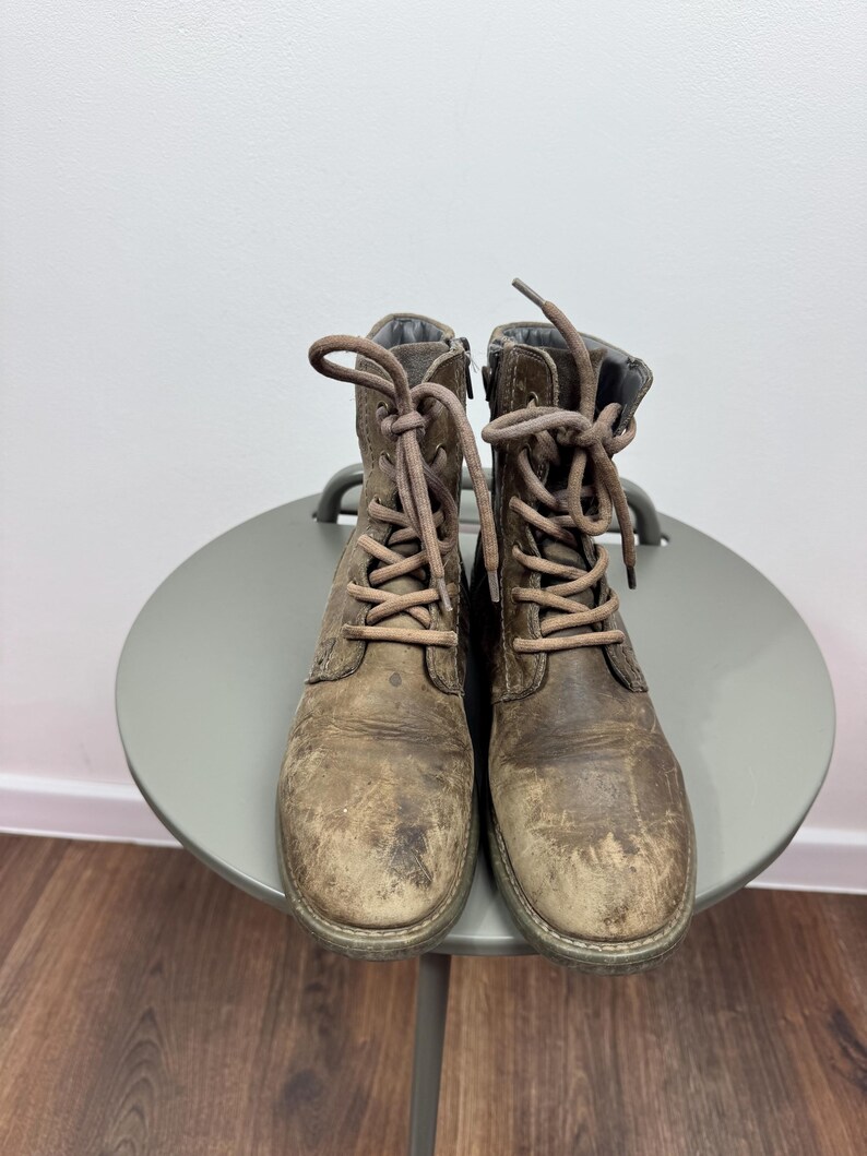 K&ouml;nnte beinhalten: Ein Paar abgenutzte braune Schn&uuml;rstiefel aus Leder mit runder Zehenpartie, passenden braunen Schn&uuml;rsenkeln und einem seitlichen Rei&szlig;verschluss. Die Stiefel weisen eine abgenutzte, strukturierte Oberfl&auml;che mit sichtbaren Kratzern auf.