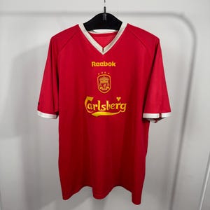 May include: A red short-sleeve jersey with a V-neck collar featuring white trim. The chest displays the yellow Reebok logo, a yellow Liverpool Football Club crest with four stars above it, and the yellow Carlsberg text logo centered below.
