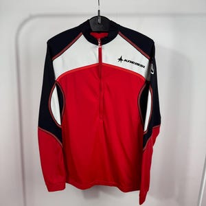 Op de afbeelding: Een sportshirt met lange mouwen en een korte rits, voorzien van een kleurblokontwerp in rood, wit en zwart. Op de borst staat de zwarte tekst KNEISSL met een sterpictogram. Het kledingstuk heeft een hoge kraag en contrasterende stiksels.