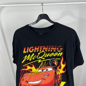 Peut inclure: T-shirt noir avec un motif de Flash McQueen du film Disney Pixar Cars. Le graphisme comprend le texte "Lightning McQueen" en jaune et orange, le num&eacute;ro "95" et la phrase "Rule the Champ".