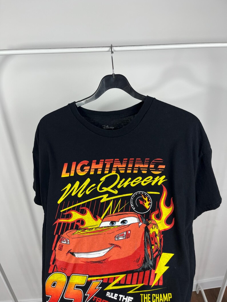 Peut inclure: T-shirt noir avec un motif de Flash McQueen du film Disney Pixar Cars. Le graphisme comprend le texte "Lightning McQueen" en jaune et orange, le num&eacute;ro "95" et la phrase "Rule the Champ".