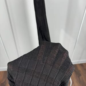 May include: A black, textured knit dress is displayed on a hanger. The fabric has a ribbed pattern, and the dress appears to be lightweight. The image is taken indoors, with a wooden floor and a white background.