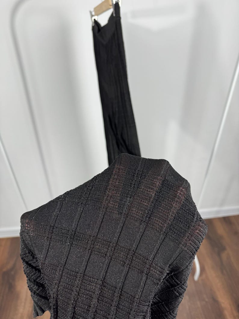 May include: A black, textured knit dress is displayed on a hanger. The fabric has a ribbed pattern, and the dress appears to be lightweight. The image is taken indoors, with a wooden floor and a white background.