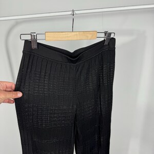 May include: Black, textured pants hanging on a wooden hanger. The pants have a high waist and a vertical ribbed pattern. The fabric appears to have a subtle sheen. The pants are displayed against a plain white background.