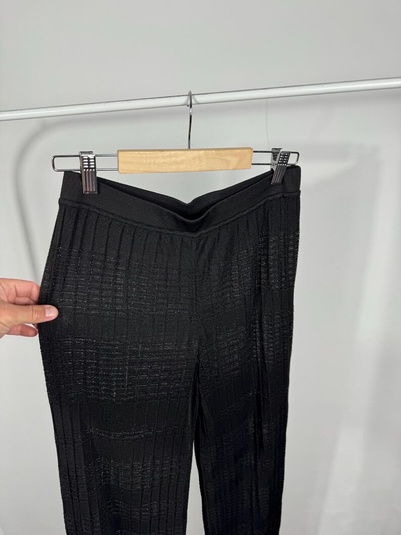 May include: Black, textured pants hanging on a wooden hanger. The pants have a high waist and a vertical ribbed pattern. The fabric appears to have a subtle sheen. The pants are displayed against a plain white background.