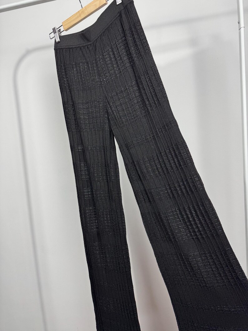 May include: Black, wide-leg pants with a textured, pleated design. The pants are hanging on a hanger against a white background. The fabric appears to have a subtle sheen.