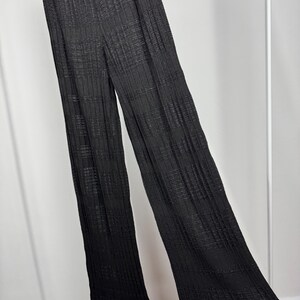 May include: Black, wide-leg pants with a textured, ribbed design. The pants are hanging on a white clothing rack. The fabric appears to be lightweight and flowing.