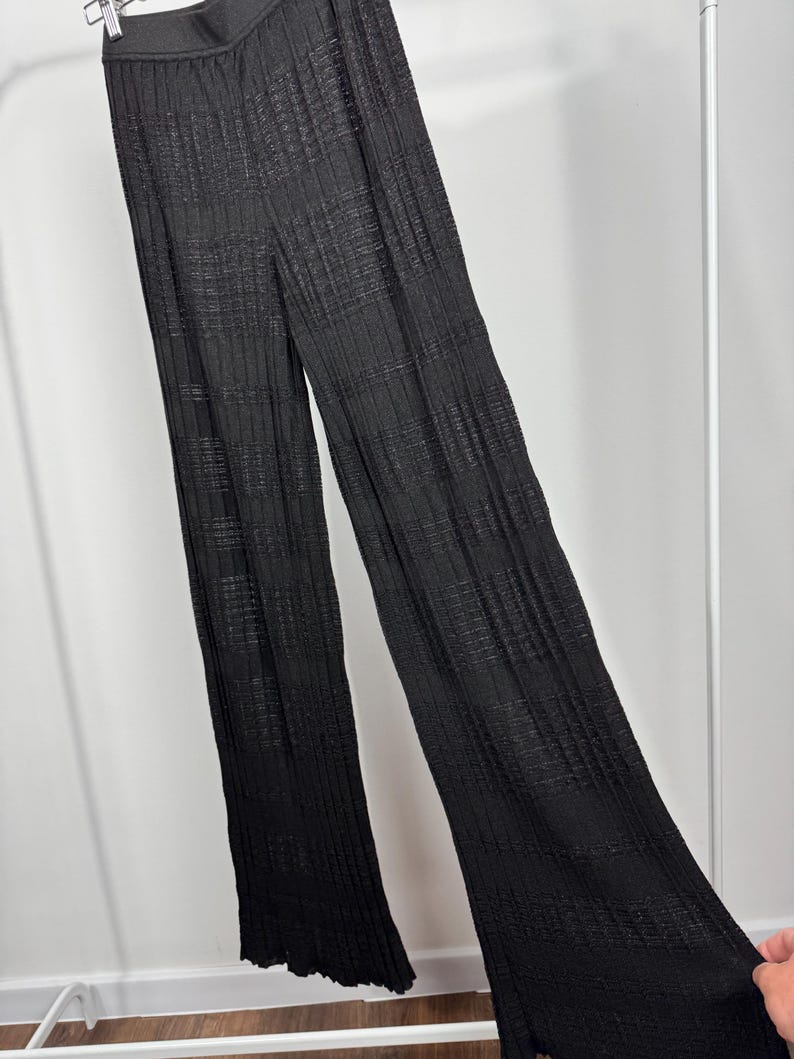 May include: Black, wide-leg pants with a textured, ribbed design. The pants are hanging on a white clothing rack. The fabric appears to be lightweight and flowing.