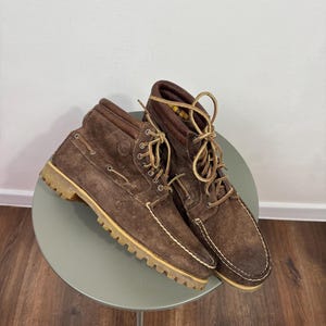 May include: A pair of dark brown suede ankle boots featuring a moccasin-style toe, light-coloured thick lug soles, and tan laces. The boots have a padded collar, metal eyelets, and a yellow and black plaid interior lining visible at the top opening.