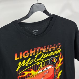 Peut inclure: T-shirt noir avec un motif de Flash McQueen du film Cars de Disney. Le graphisme comprend le texte "Lightning McQueen" en jaune et orange, une voiture de course rouge et des flammes. Le t-shirt a un col rond et un logo Disney.
