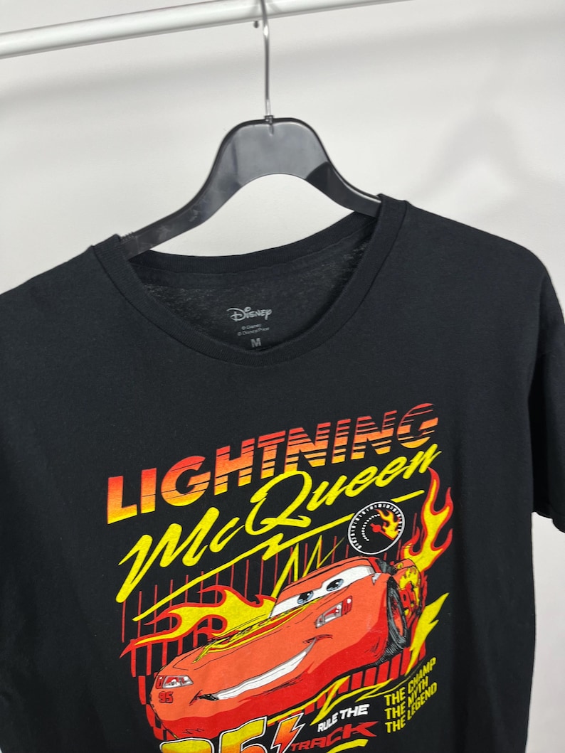 Peut inclure: T-shirt noir avec un motif de Flash McQueen du film Cars de Disney. Le graphisme comprend le texte "Lightning McQueen" en jaune et orange, une voiture de course rouge et des flammes. Le t-shirt a un col rond et un logo Disney.