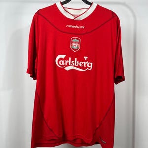 May include: A red short-sleeved soccer jersey featuring a white collar, the Reebok logo, a Liverpool Football Club crest, and the Carlsberg brand name printed in white across the chest. The shirt has dark stitching details and a loose fit.
