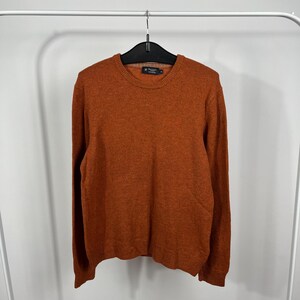 Puede incluir: Un suéter de cuello redondo color naranja quemado con mangas largas. El suéter está colgado de una percha negra sobre un fondo blanco. Una pequeña etiqueta es visible cerca del cuello.