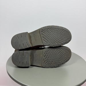 K&ouml;nnte beinhalten: Ein Paar braune Schuhe mit dunkelgrauen Gummisohlen, die ein strukturiertes Rautenmuster und einen gerillten Absatz aufweisen. Das Wort Clarks ist in der Mitte jeder Sohle eingepr&auml;gt. Die Schuhe sind &uuml;bereinander gestapelt und zeigen die Unterseite.