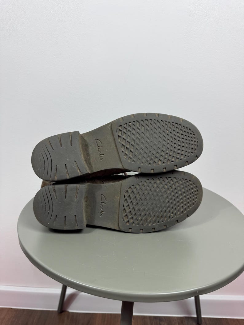 K&ouml;nnte beinhalten: Ein Paar braune Schuhe mit dunkelgrauen Gummisohlen, die ein strukturiertes Rautenmuster und einen gerillten Absatz aufweisen. Das Wort Clarks ist in der Mitte jeder Sohle eingepr&auml;gt. Die Schuhe sind &uuml;bereinander gestapelt und zeigen die Unterseite.