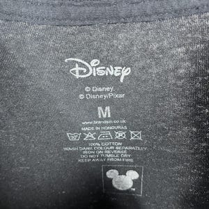Peut inclure: Gros plan sur une &eacute;tiquette de t-shirt noire avec le logo Disney et les textes "Disney" et "Disney/Pixar". L'&eacute;tiquette indique &eacute;galement la taille "M", des instructions de lavage et le texte "100% Coton".